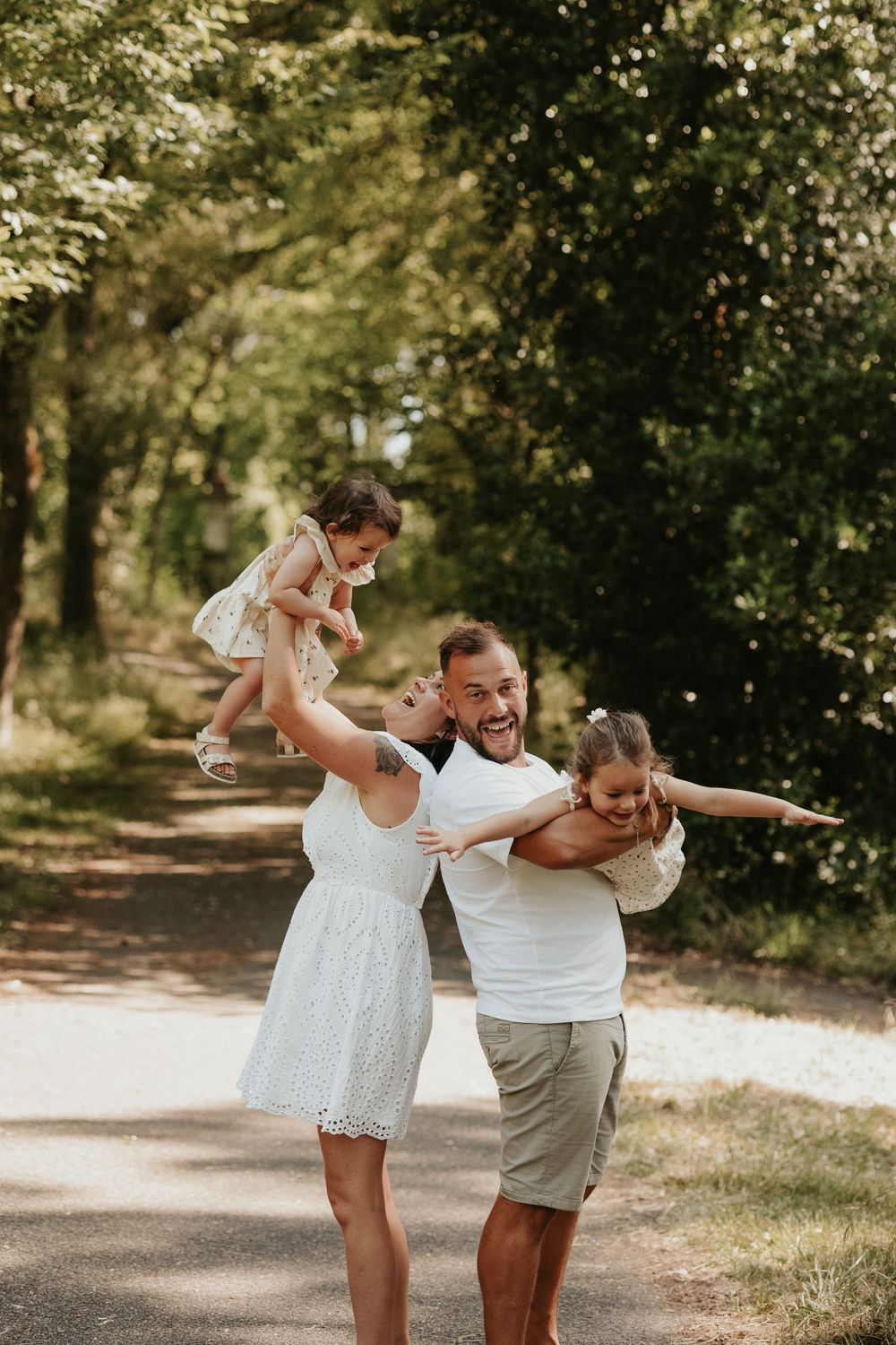 Happy family playing outdoors on a sunny park path, parents lifting two young daughters with joy and laughter.