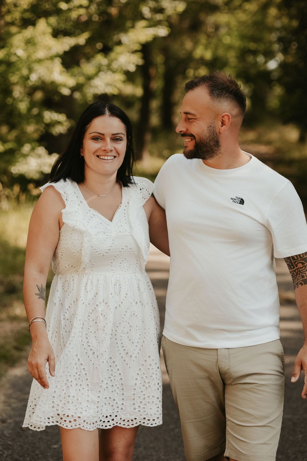 A smiling couple walks together outdoors, woman in white dress and man in white North Face tee, surrounded by lush green trees.