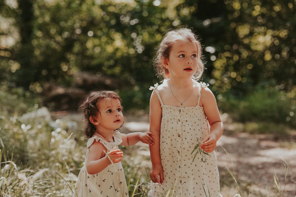 Two young girls in matching floral sundresses stand together outdoors in a sunlit garden setting.