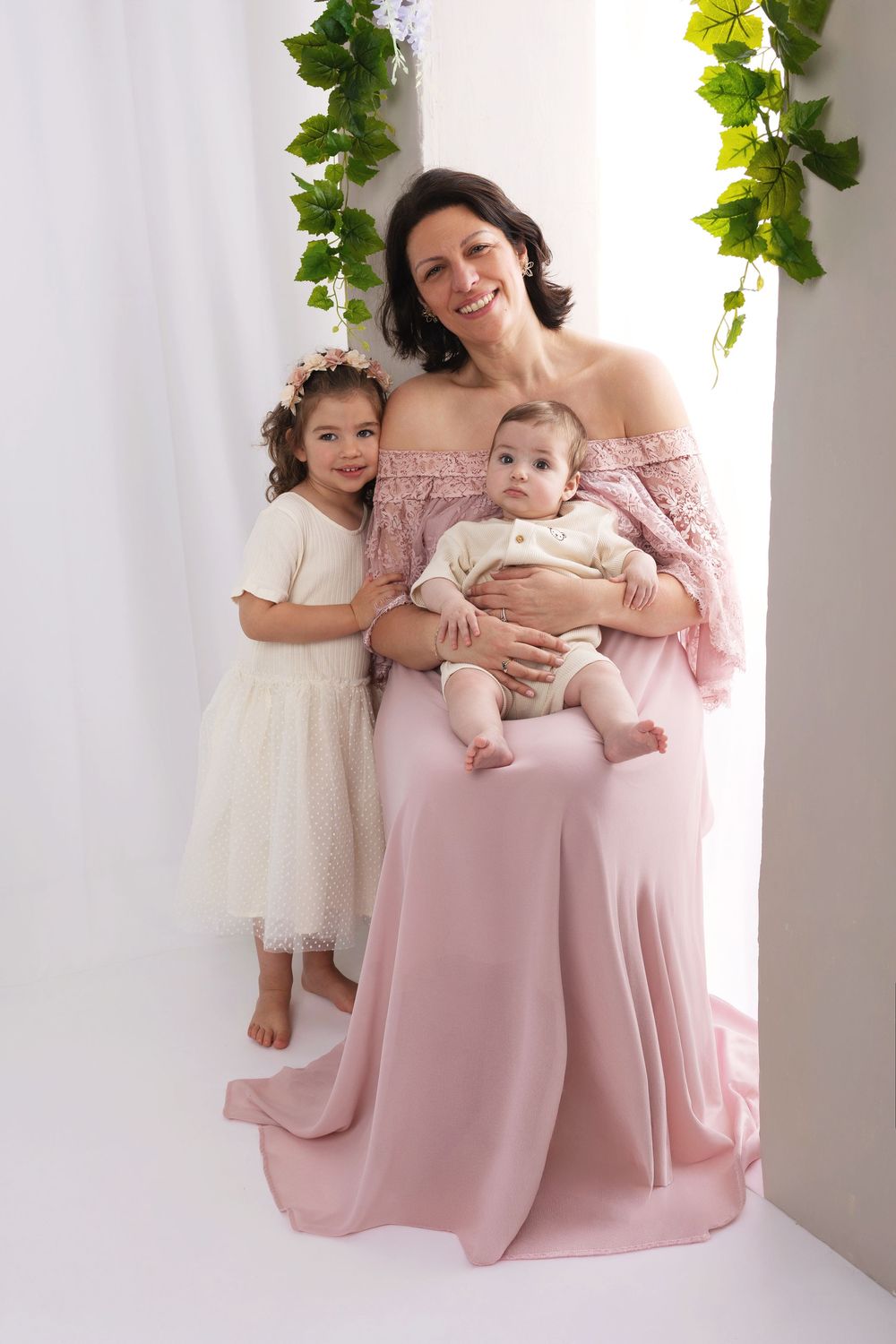 A smiling mother in a pink gown holds her baby while her young daughter in white dress stands beside her.