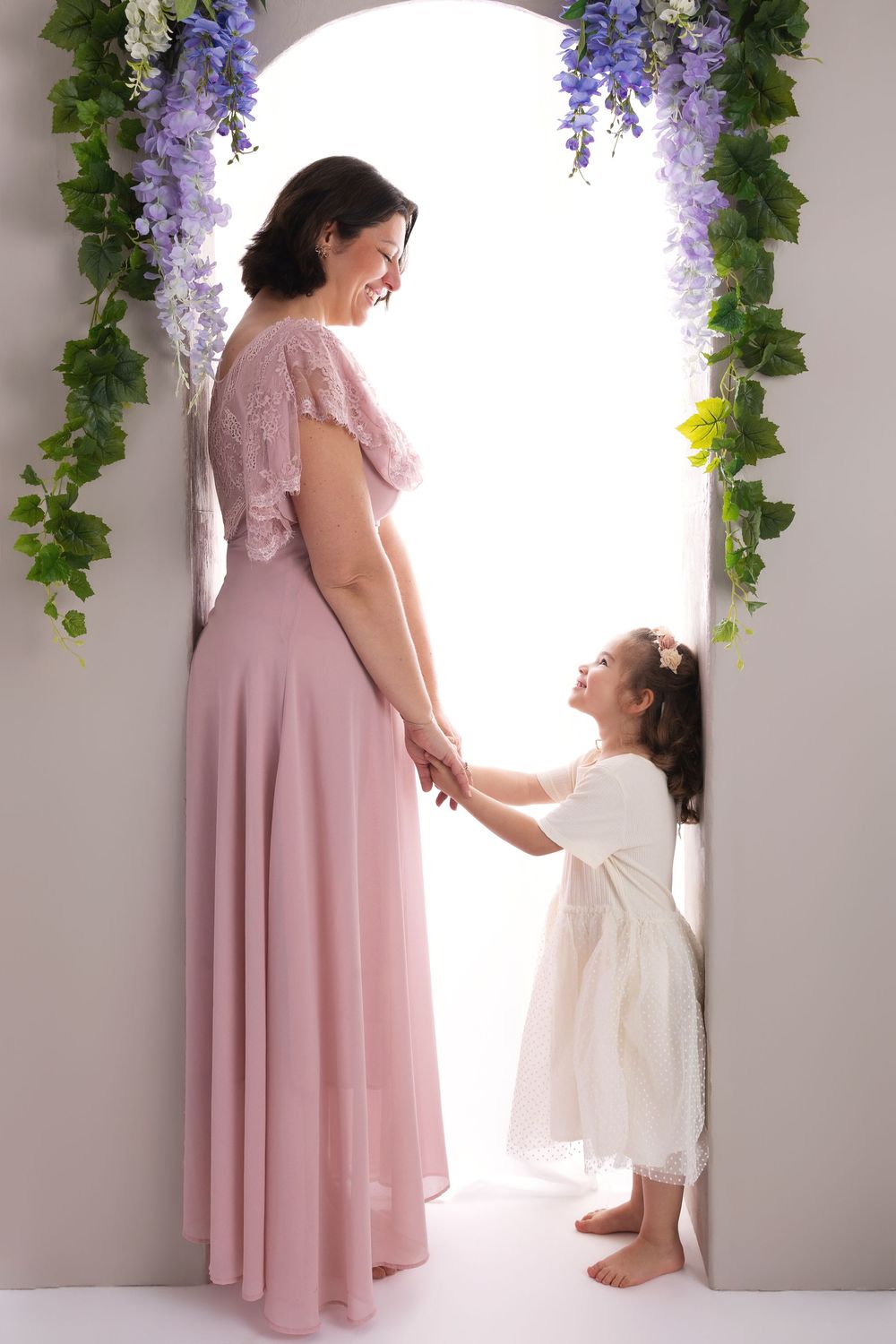 Mother in pink gown holds hands with young daughter in white dress under floral arch doorway.