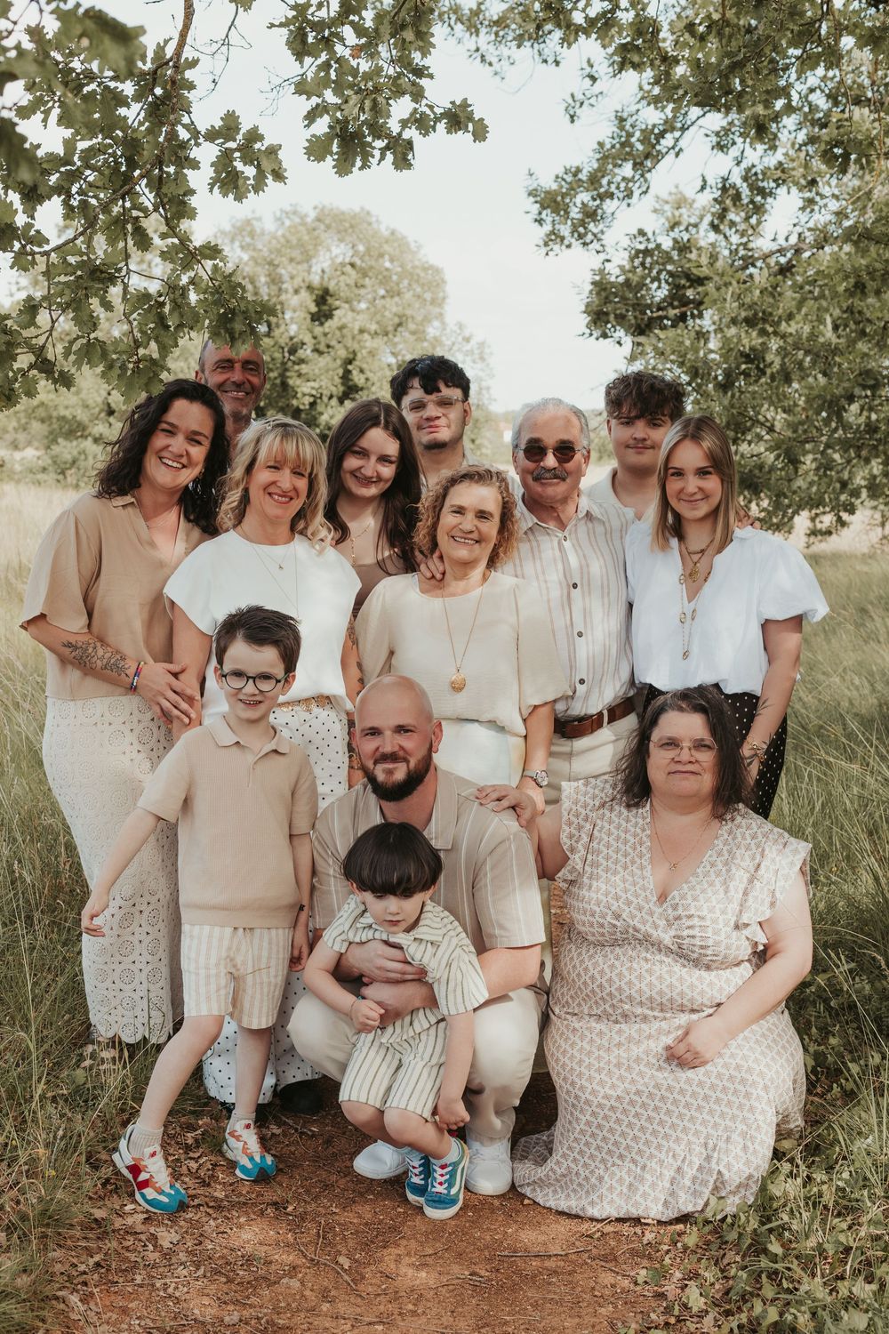 Large multigenerational family portrait outdoors in a field, dressed in neutral tones, smiling together under leafy trees.