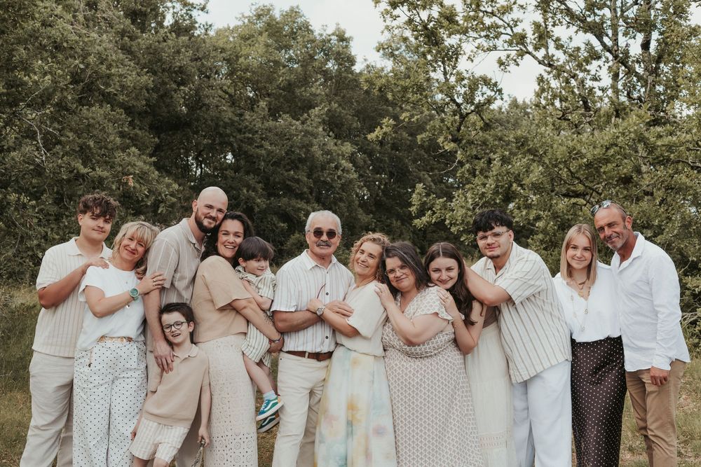 Large family group portrait outdoors in coordinated neutral outfits, smiling together against lush green trees.