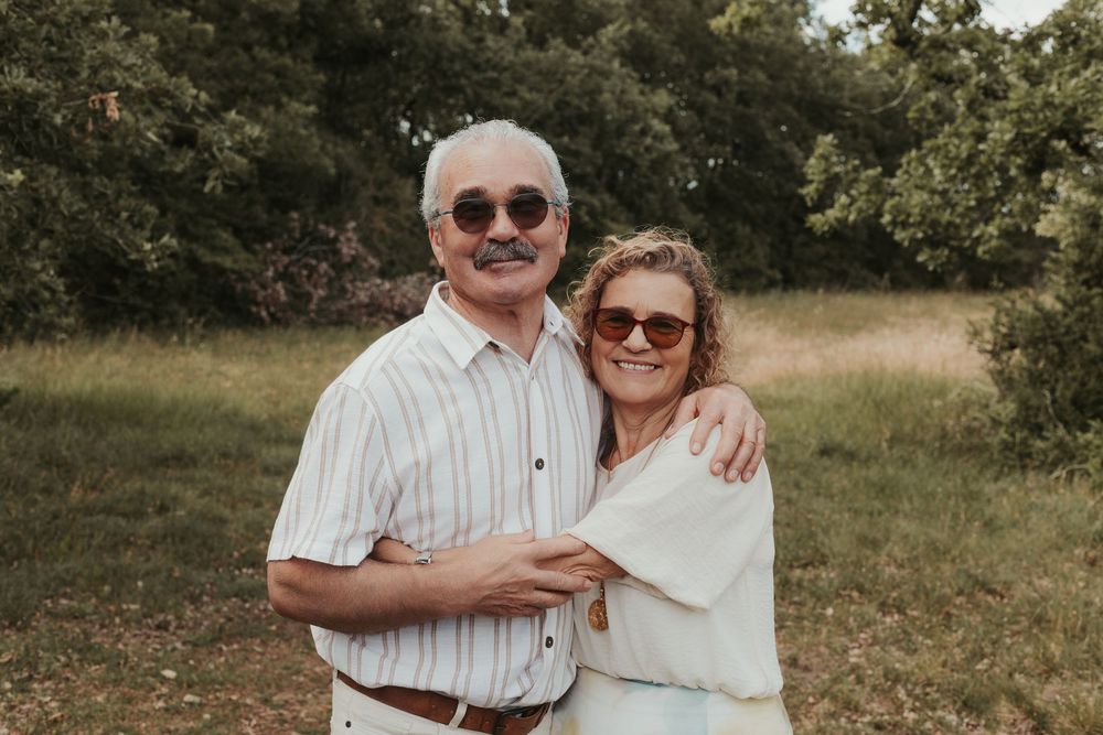 An older couple smiling outdoors, wearing sunglasses, embracing in a lush green field.