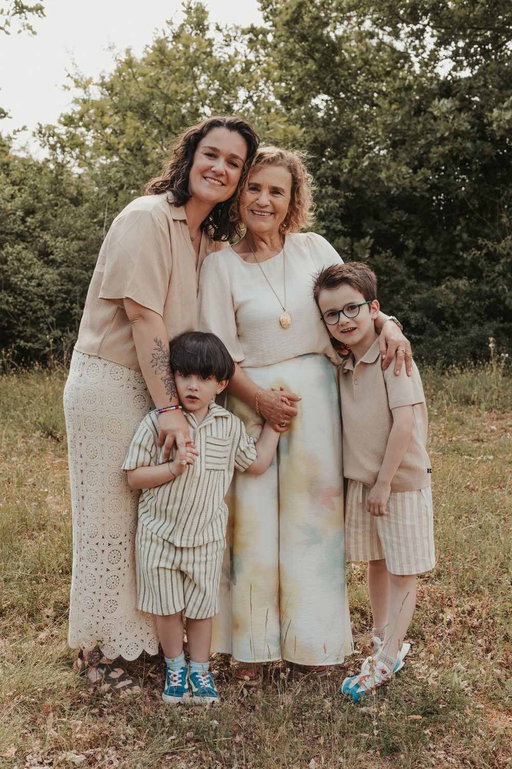 Family portrait of two women and two boys posing outdoors in coordinated neutral outfits among green trees.