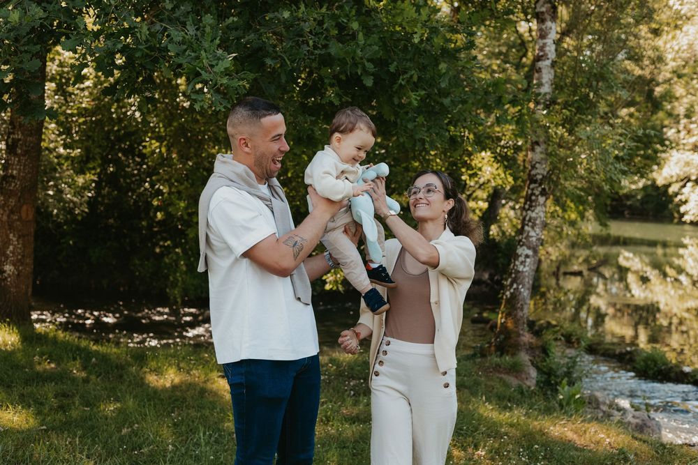 Happy family with parents lifting smiling baby outdoors in lush green park setting on a sunny day.