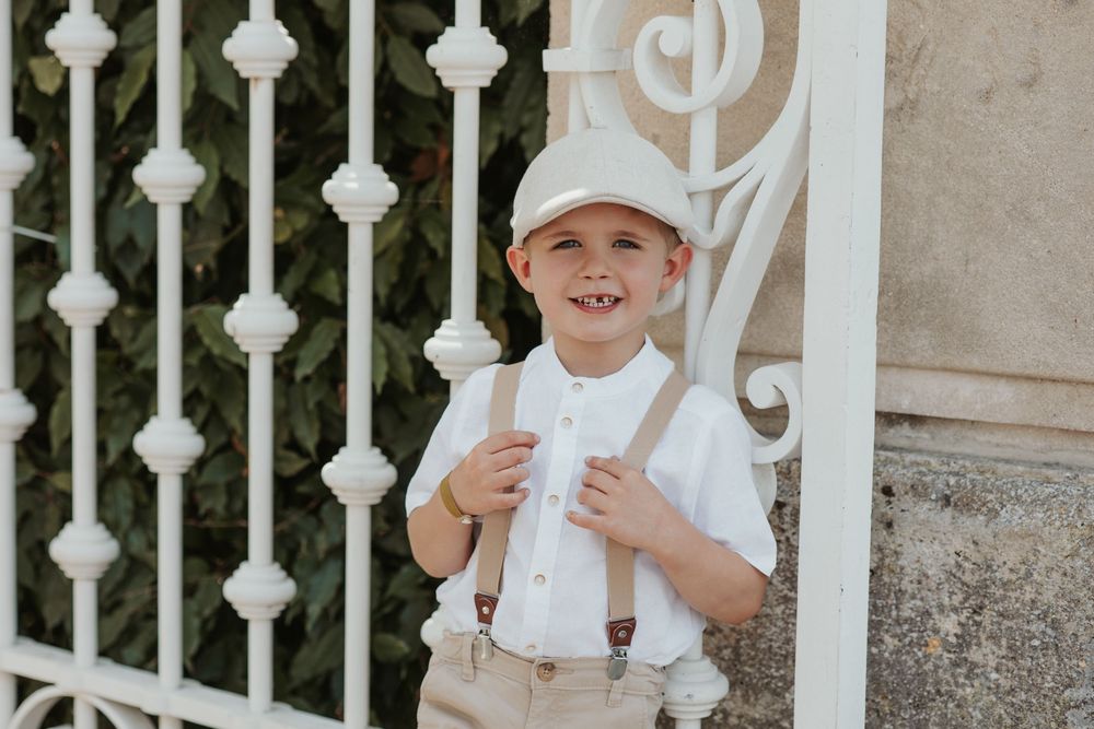 Smiling boy in white shirt, suspenders, and newsboy cap posing by ornate white iron gate.
