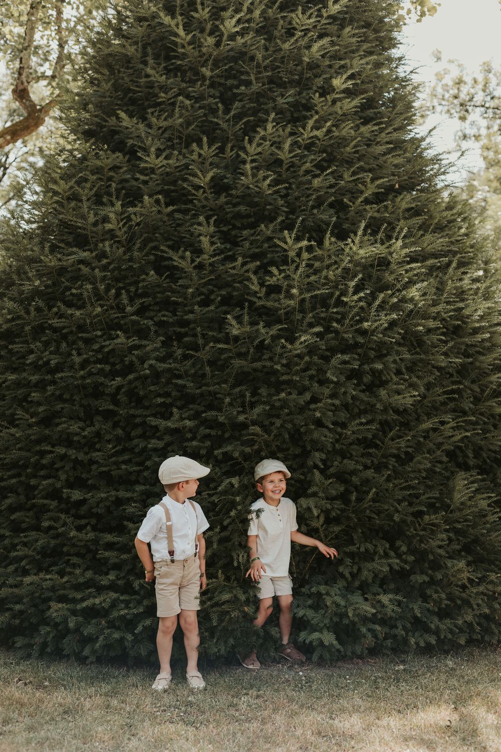 Two young boys in matching beige outfits with suspenders and flat caps standing outdoors in front of a large evergreen tree.