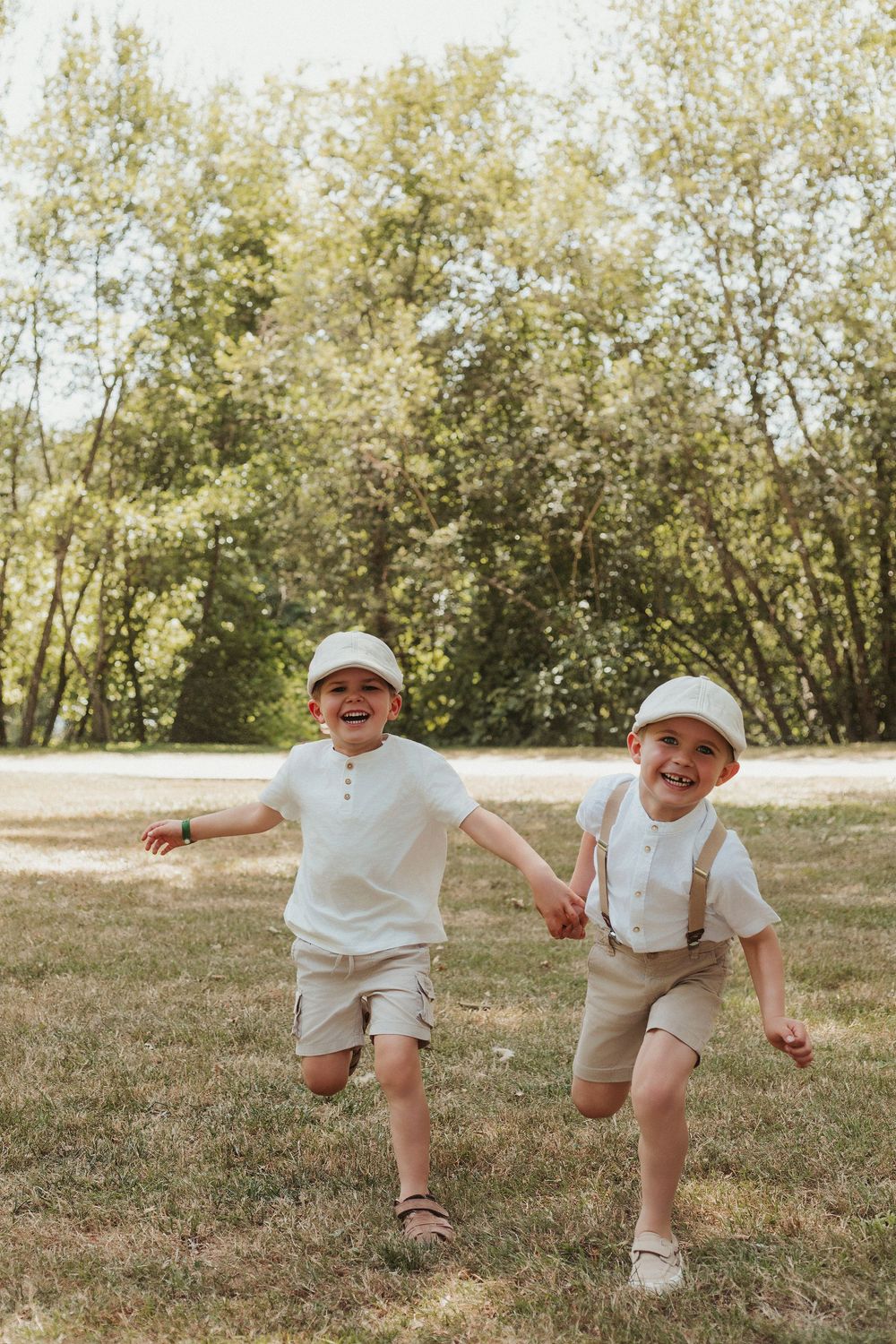 Two joyful boys in matching white outfits and caps run hand-in-hand across a sunny green park.
