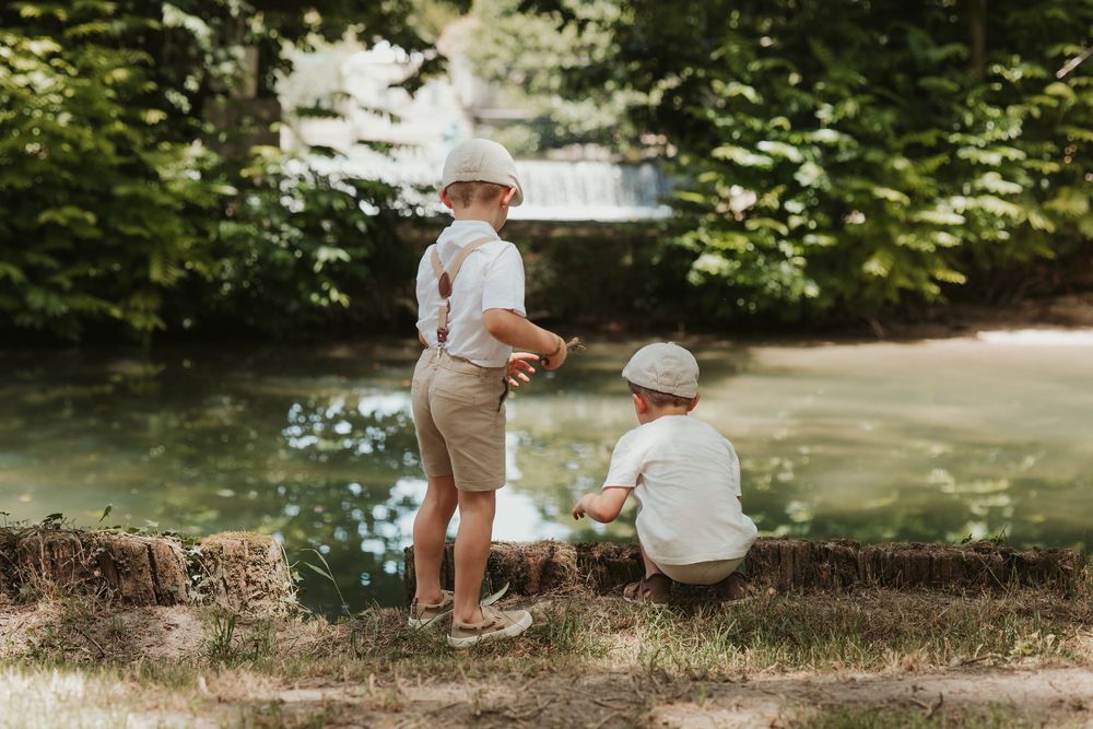 Two young boys in matching outfits and hats fishing by a serene pond surrounded by lush green trees.