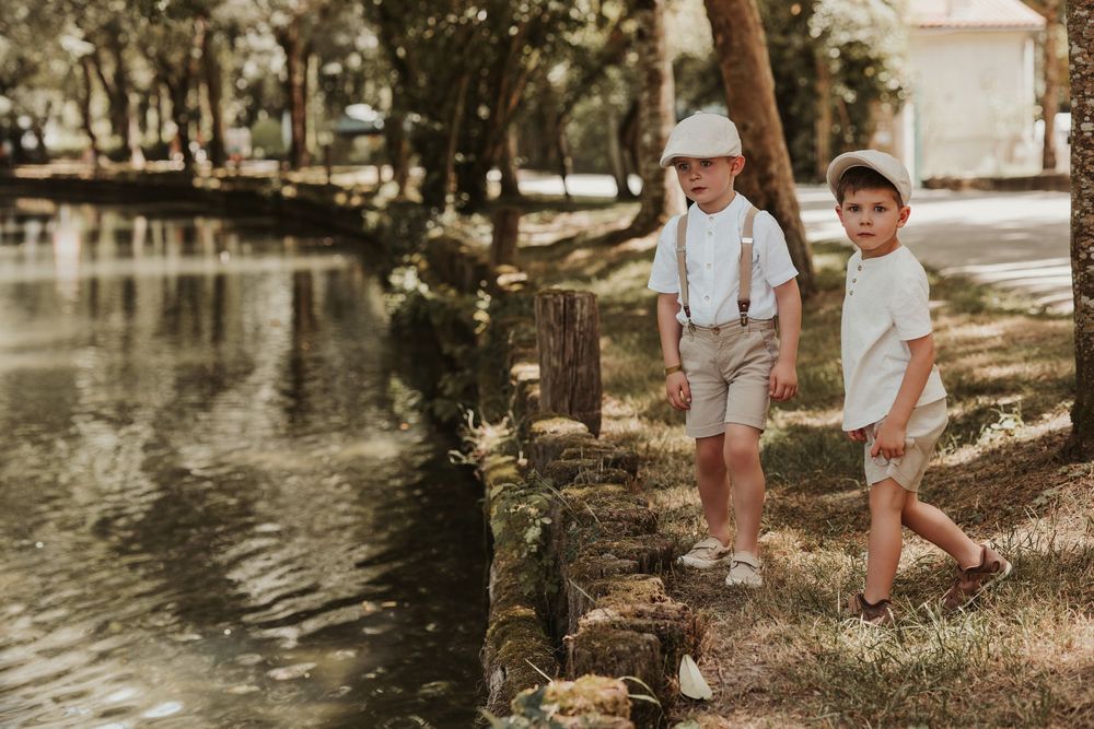 Two young boys in suspenders and caps stand by a serene pond in a lush green park.