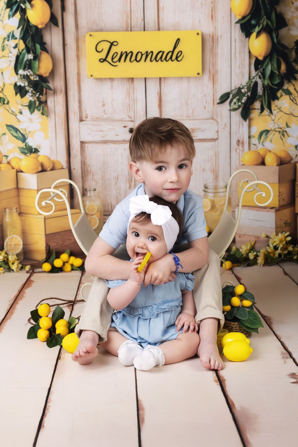 Two young siblings pose in a lemon-themed photo shoot, with a baby girl and toddler boy sitting together under a Lemonade sign.