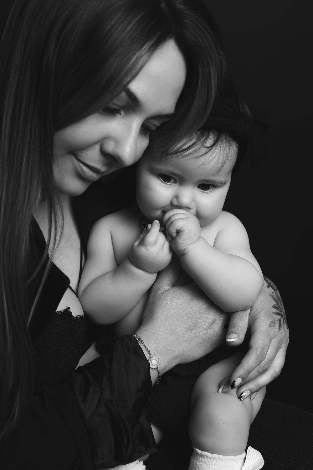 Black and white portrait of a smiling mother holding her baby, who is chewing on fingers, against dark background.