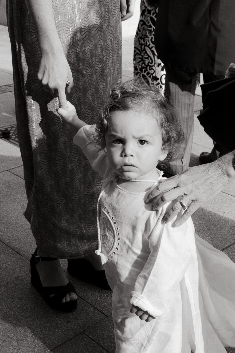 Small child in formal white attire stands holding an adult's hand at a formal event.
