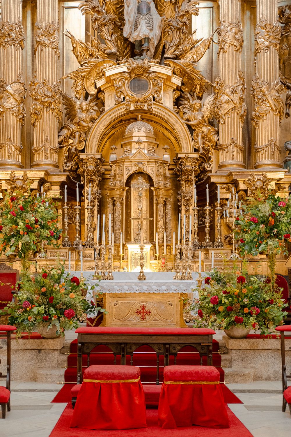 Ornate gold altar with red kneeling cushions and floral arrangements in a baroque church interior.