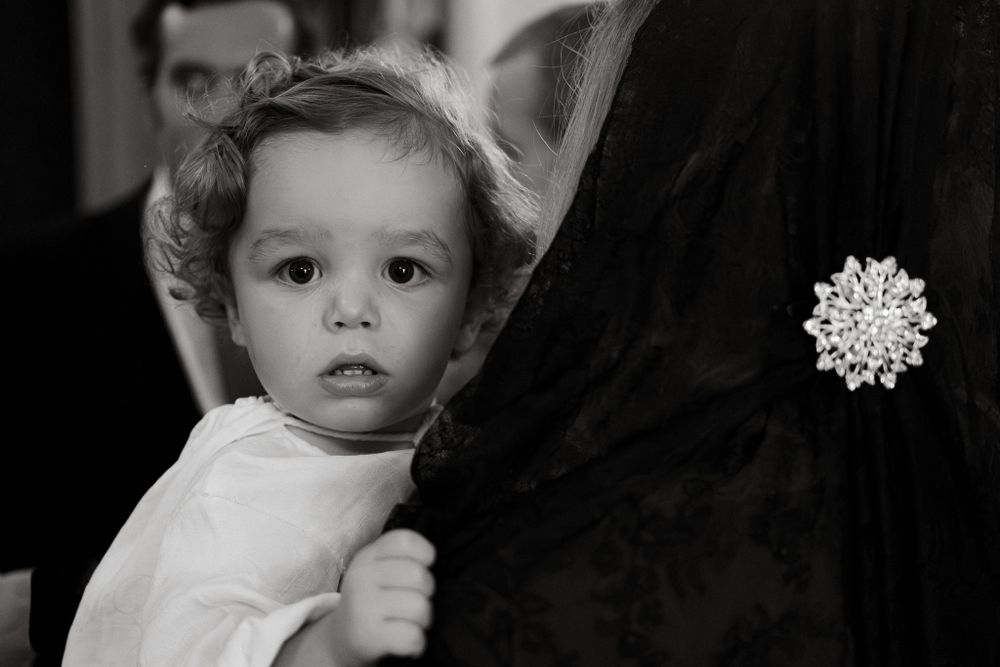 A young child in white clothing holds onto a dark garment while looking at the camera.