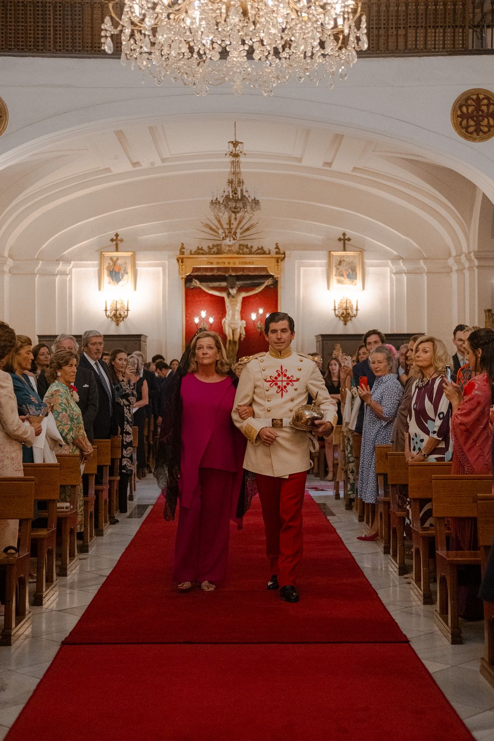 Formal church ceremony with attendees in formal attire walking down red-carpeted aisle.