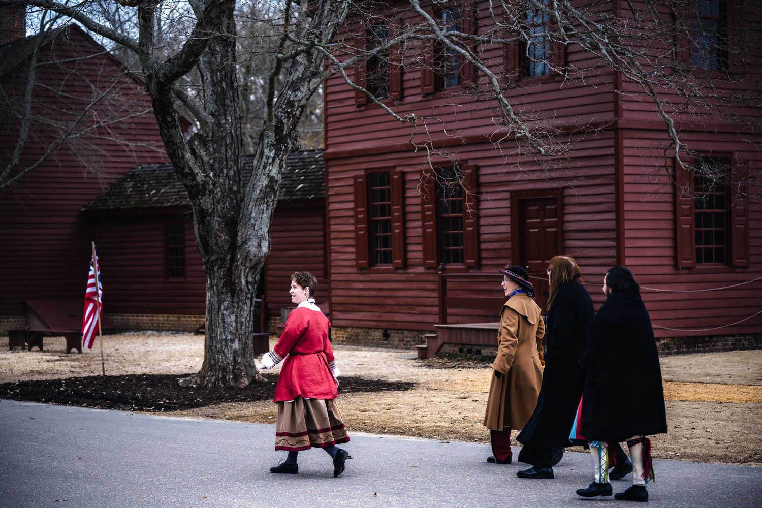 Historic colonial era building with figures in period dress walking past an American flag on a winter day.