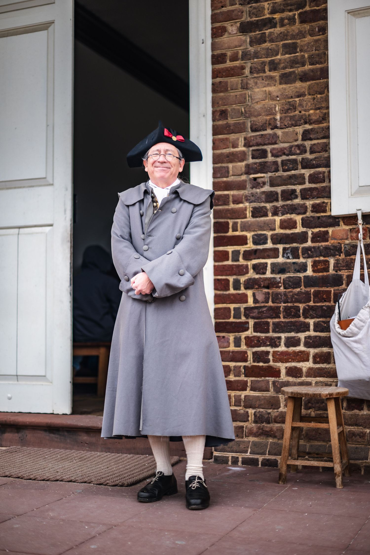 A person in colonial style clothing wearing a tricorn hat and gray coat stands in a brick doorway.