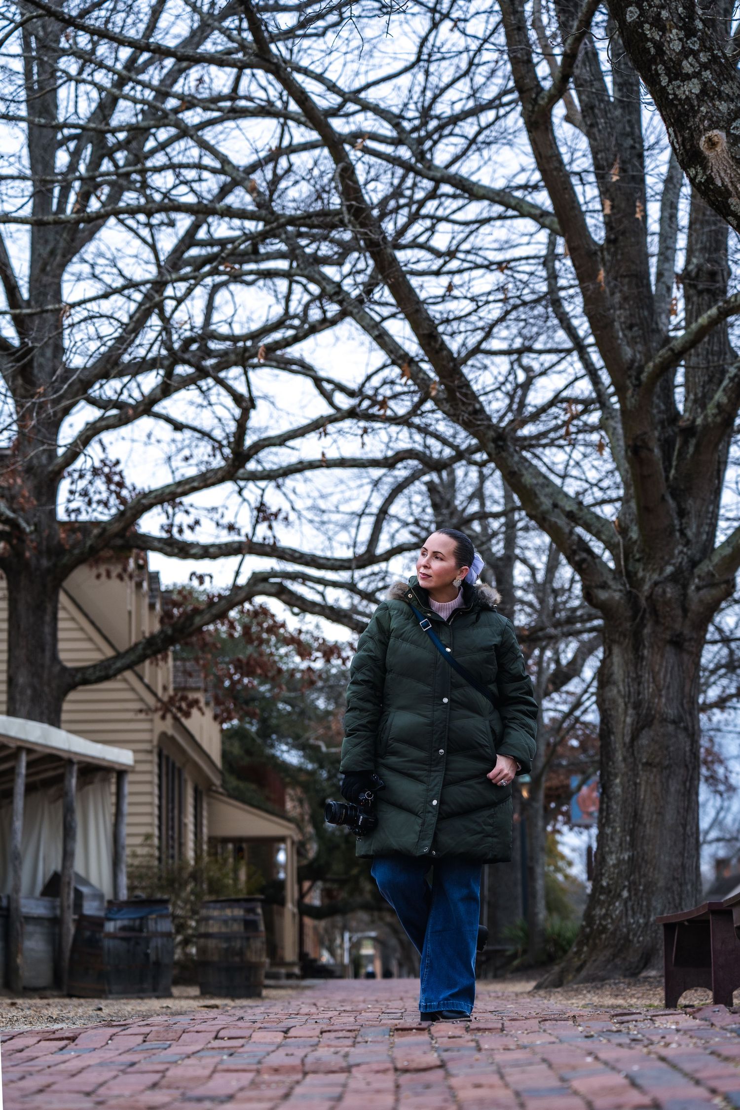 Person walking along a brick pathway surrounded by bare winter trees and a tan house in the background on an overcast day.