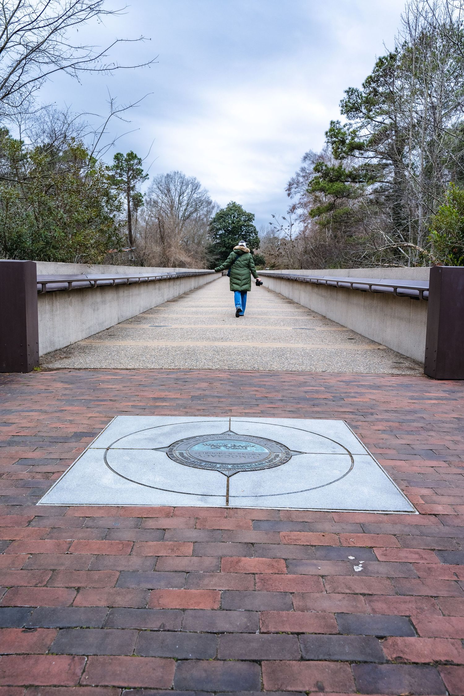 A person in a green jacket walks along a long concrete bridge with red brick paving and a compass design at the entrance.