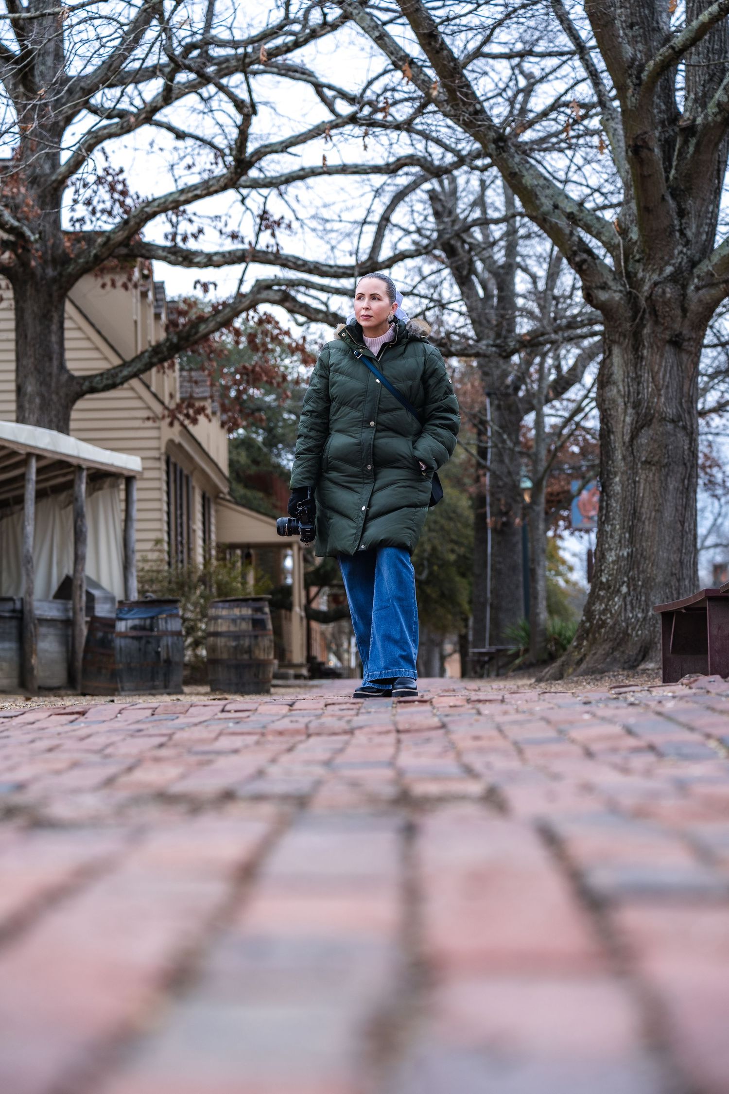 Person in green winter coat and blue pants standing on a brick pathway during autumn with trees in the background.