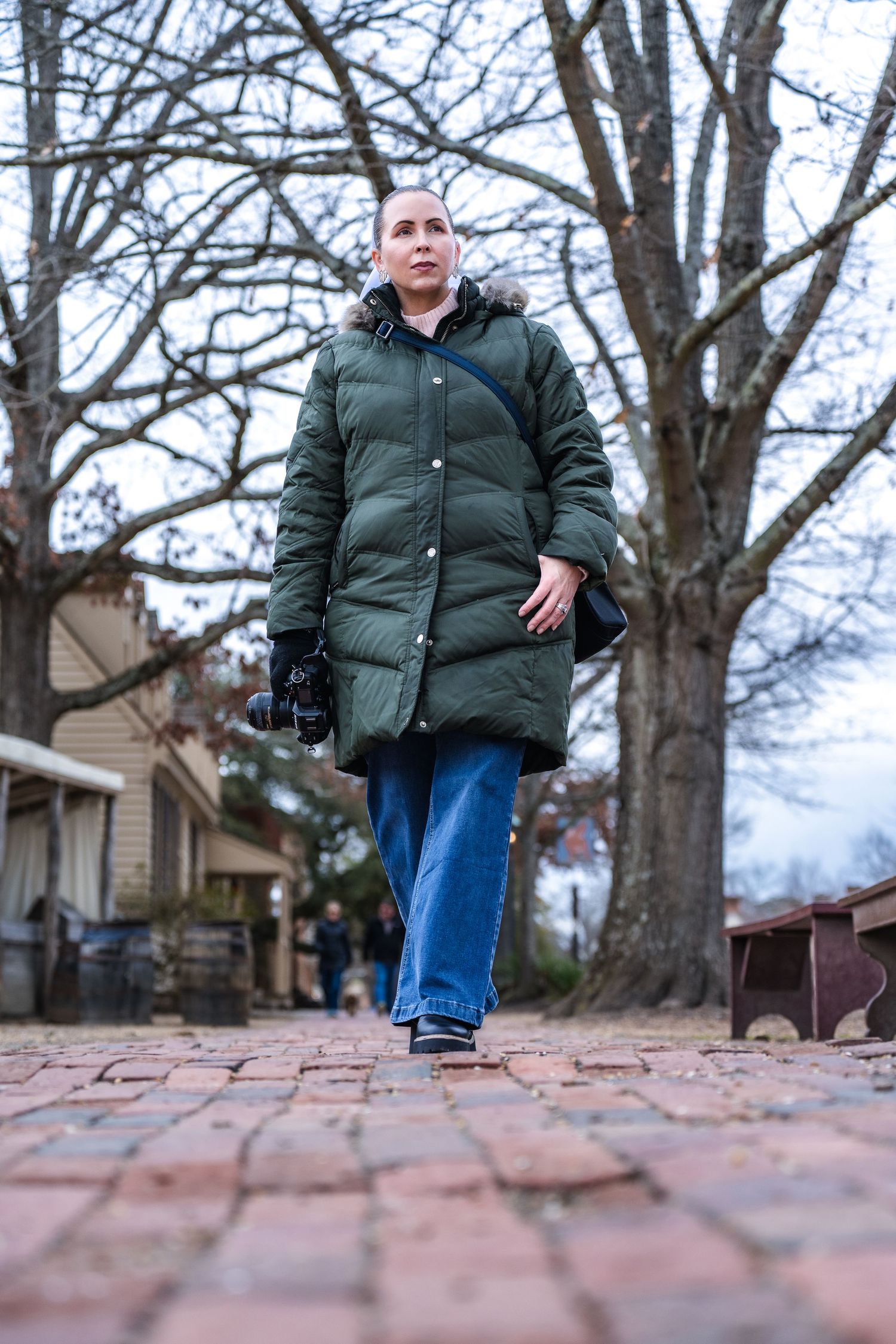 Person wearing a green puffer coat and jeans walking along a brick path on a winter day with barren trees in background.