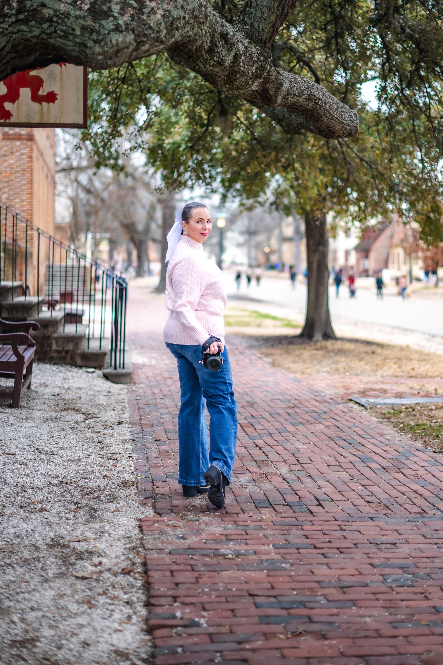 Person in pink sweater and jeans standing on a brick sidewalk path bordered by trees in a historic neighborhood.