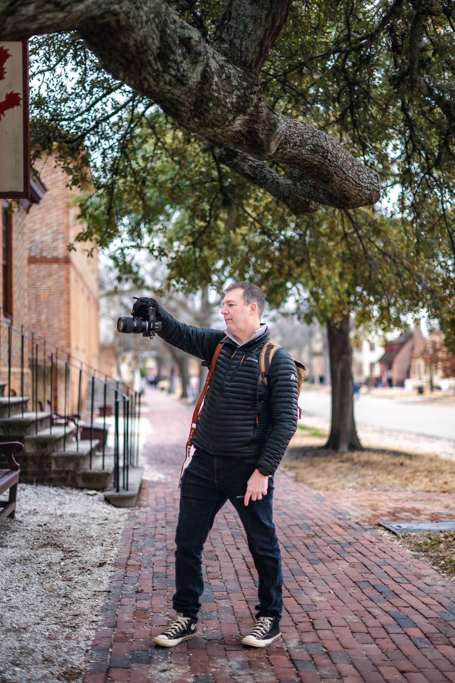 Person in black winter attire stands on brick sidewalk near large tree and historic buildings in urban setting.