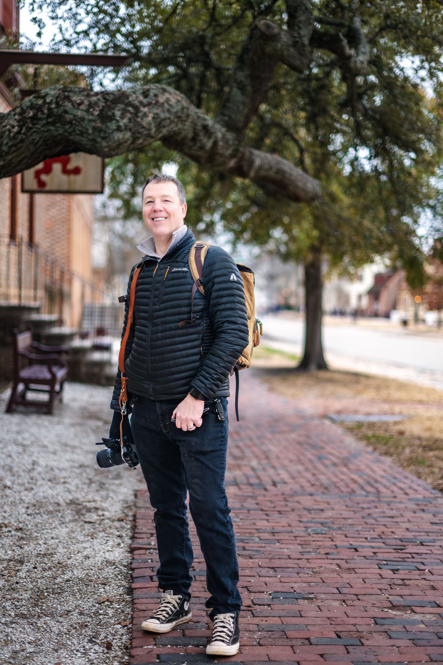 A photographer with camera gear and backpack stands smiling on a brick pathway beneath a curved tree branch.