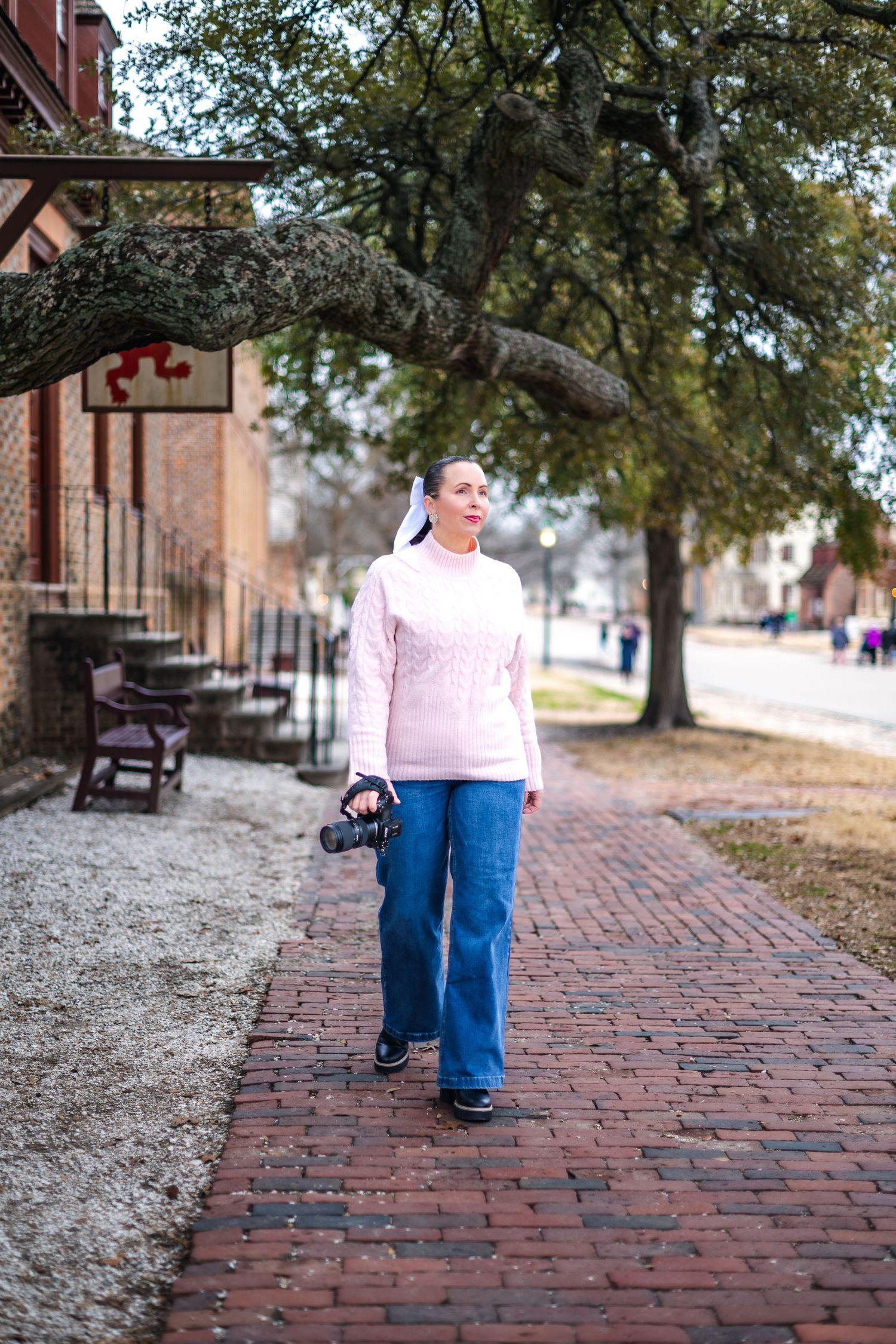 Photographer with camera walking on brick sidewalk past historic buildings on tree-lined street.