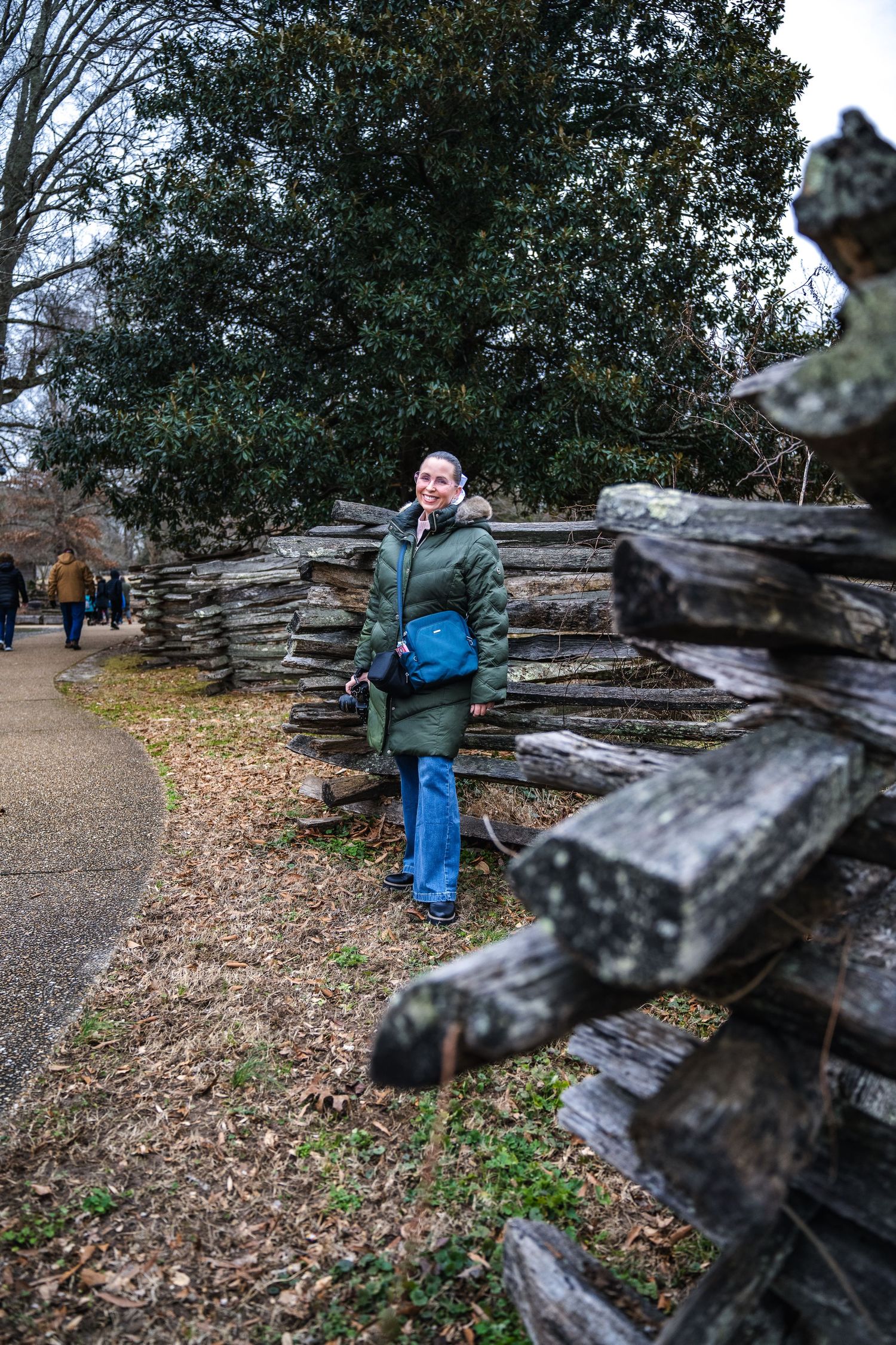 Person in green jacket and jeans stands beside rustic split rail fence on autumn path with fallen leaves.