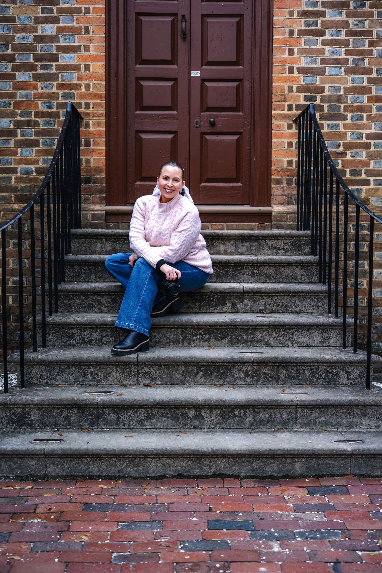 Someone in a pink puffy jacket sits on concrete steps in front of a brown door and brick building.