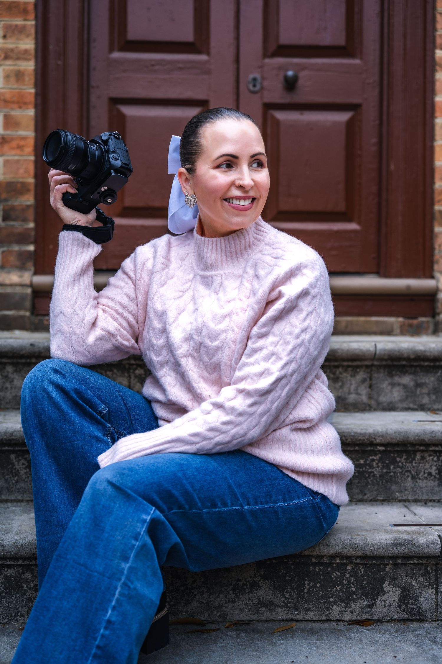 Photographer in pink cable knit sweater and jeans sits on steps while holding a DSLR camera and smiling.