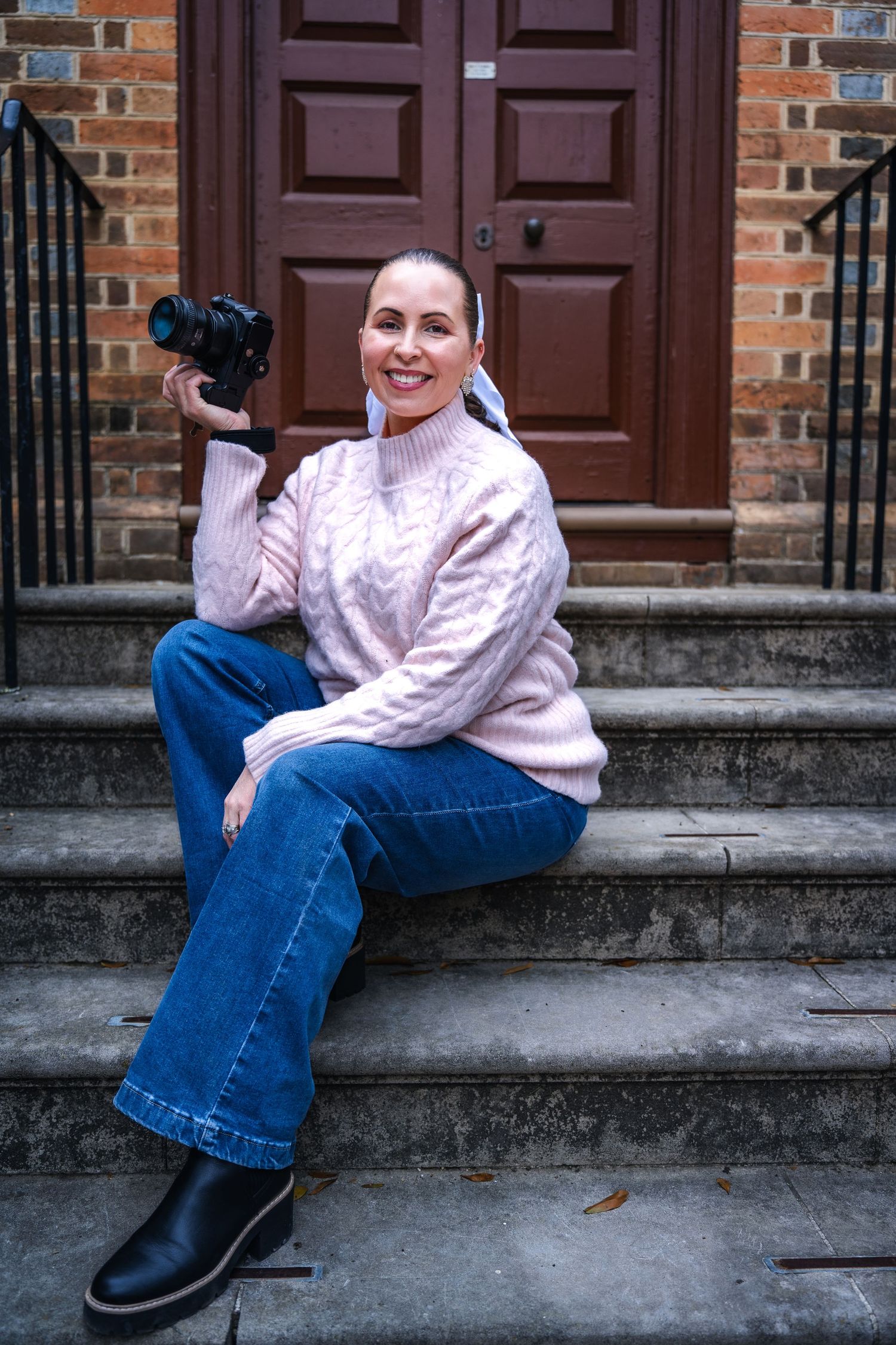 Photographer in cozy pink sweater and jeans sits on stone steps holding camera by brown wooden door.