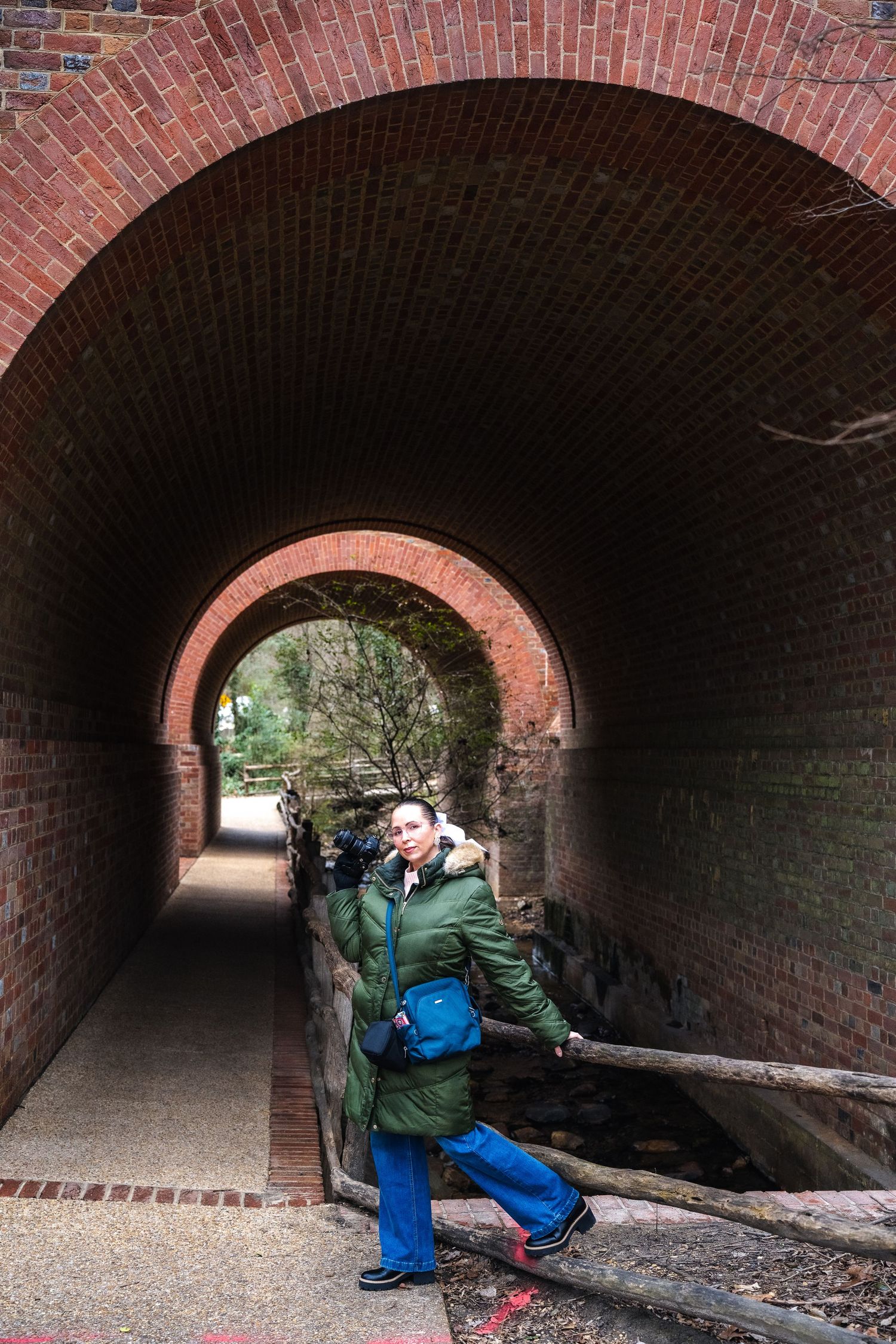 A brick tunnel walkway in a park with a curved archway entrance leading to a green wooded path ahead.