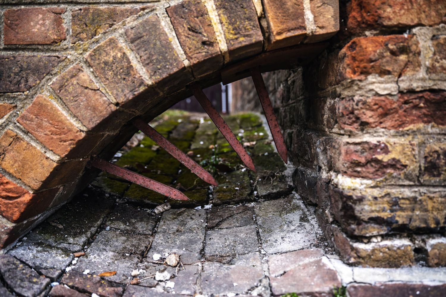 Crumbling brick archway with exposed bars reveals glimpse of greenery through weathered stone wall.