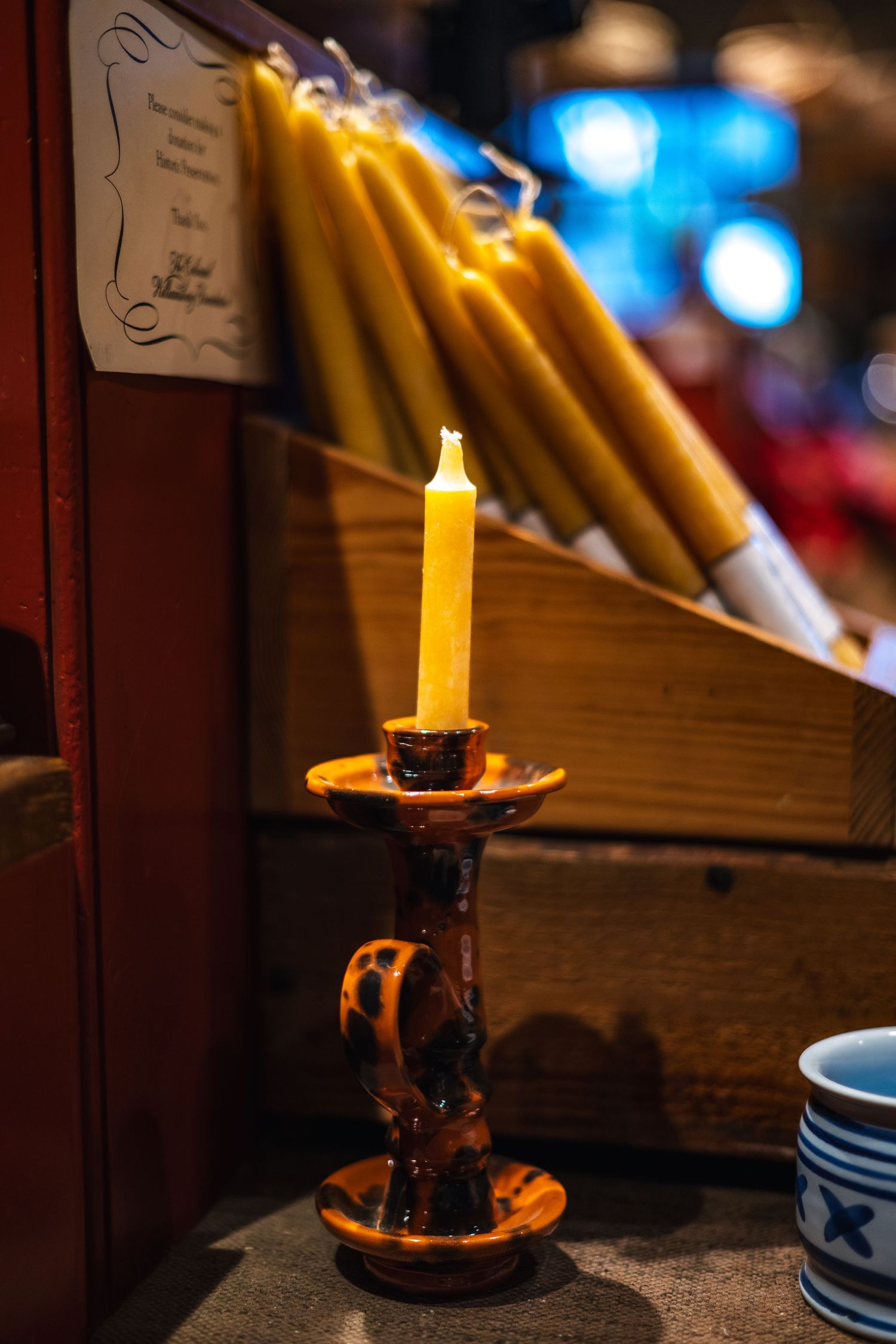 A glowing yellow candle in an antique brass candlestick holder sits near stacked taper candles with a blue bokeh background.