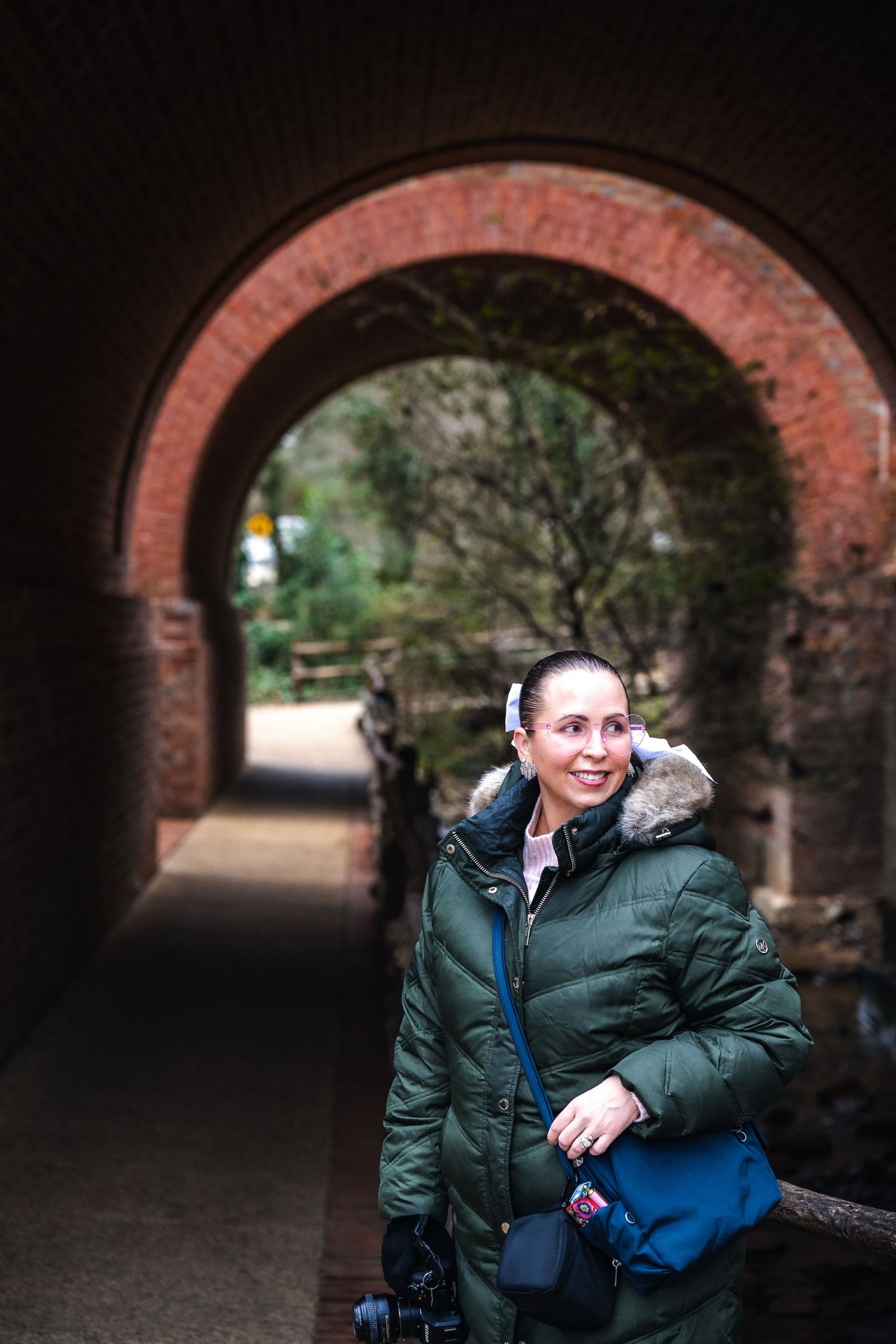Person in a green winter coat with fur hood standing beneath a red brick archway in a park.