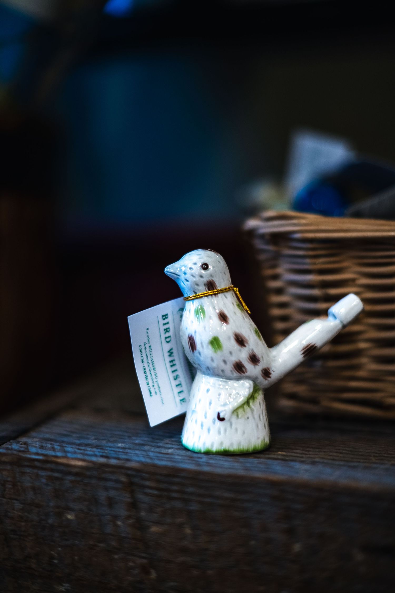 A small ceramic bird whistle with colorful polka dots sits on a dark wooden surface next to a woven basket.