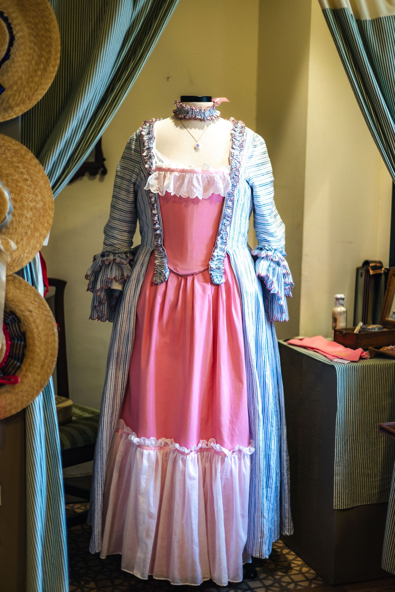 A vintage colonial style dress with pink bodice and white trim displayed on mannequin next to straw hats and blue curtains.