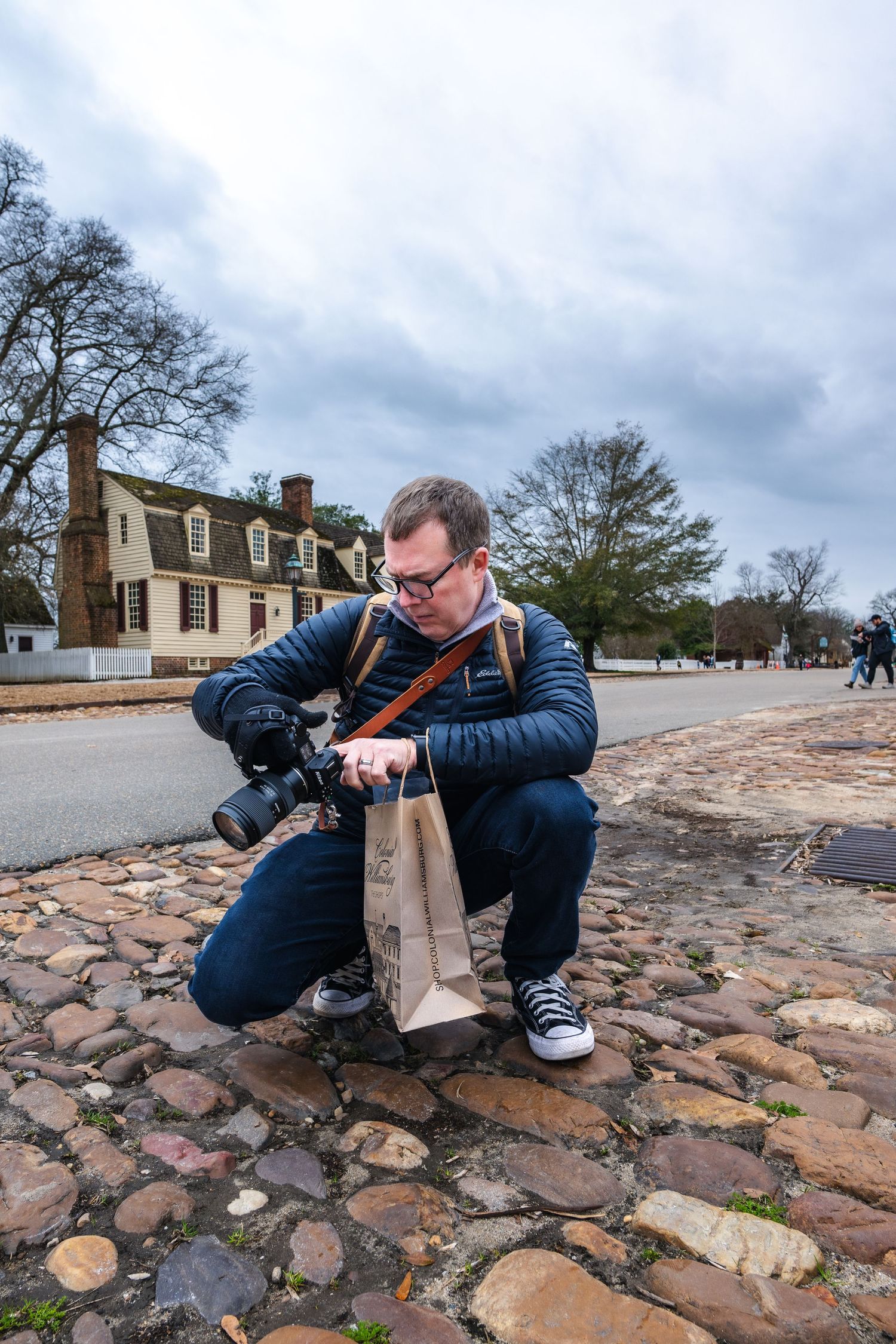 A photographer with camera gear crouches on historic cobblestone streets in Colonial Williamsburg on a cloudy day.