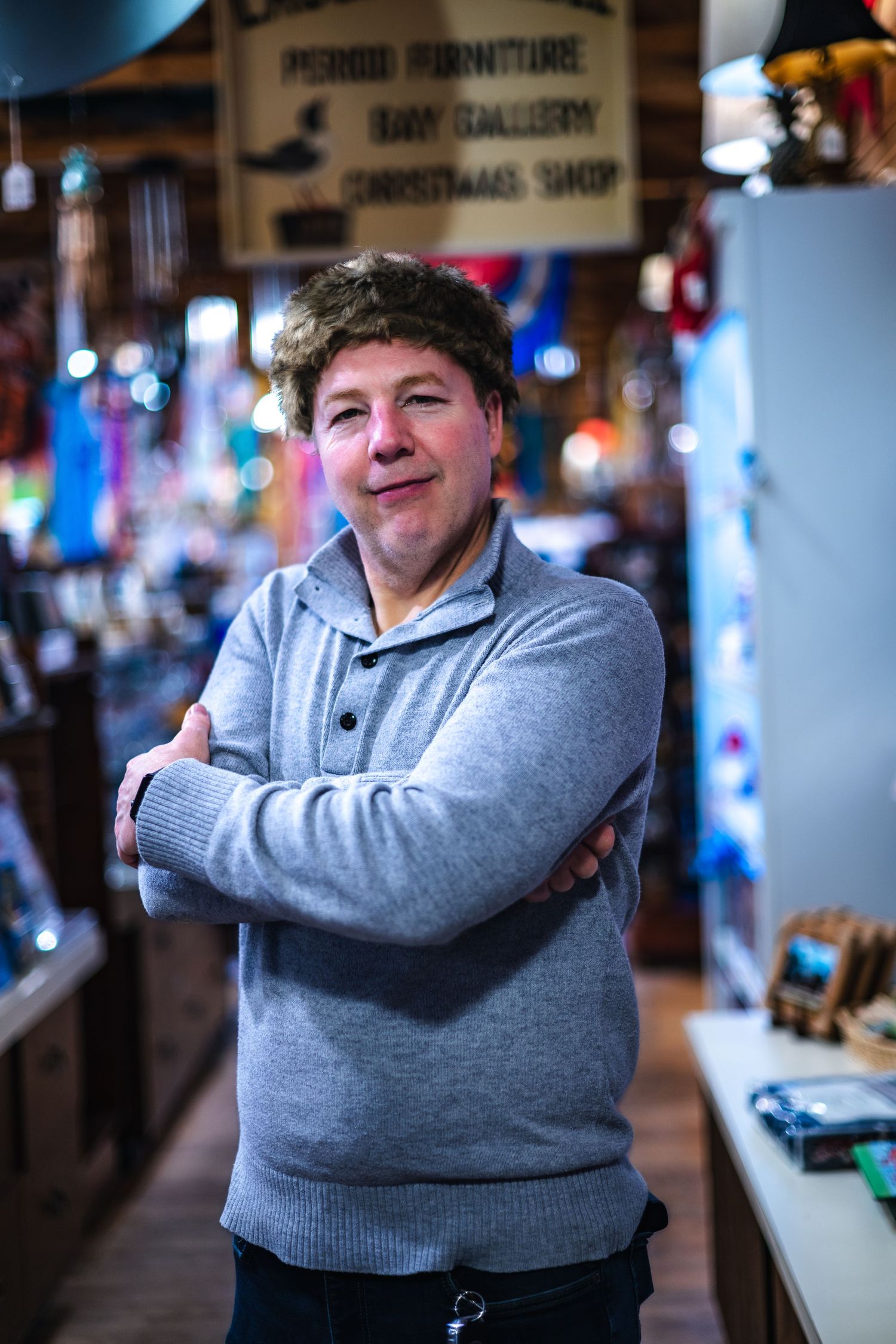 A person in a gray sweater stands with crossed arms inside what appears to be a shop or retail space in warm lighting.