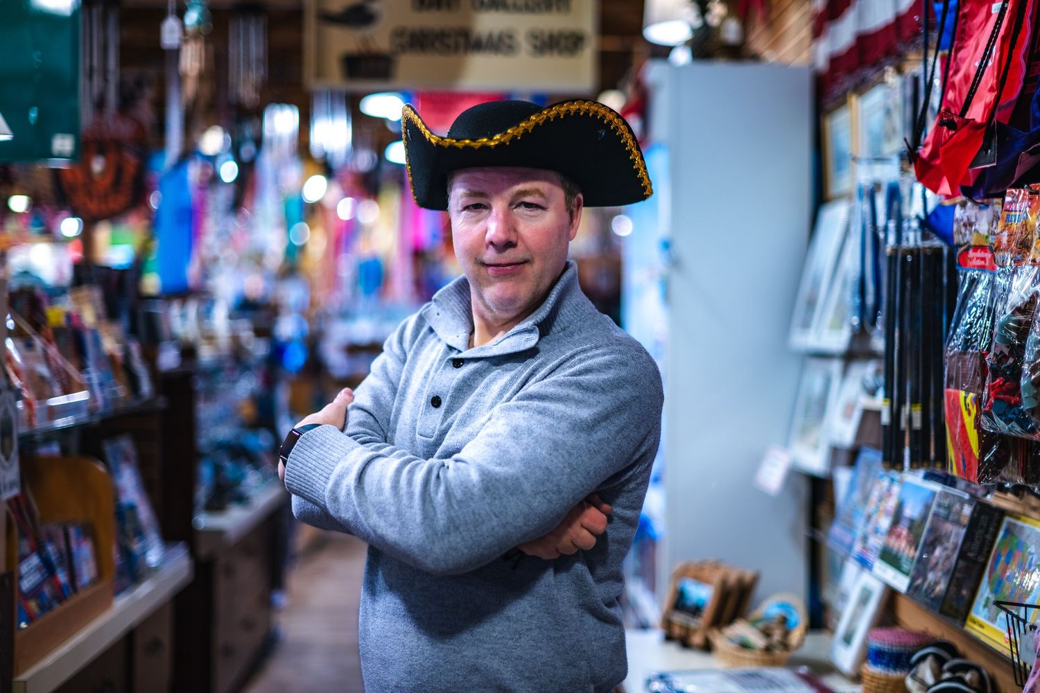 Shop vendor wearing tricorn colonial hat and gray sweater poses with crossed arms in colorful souvenir store.