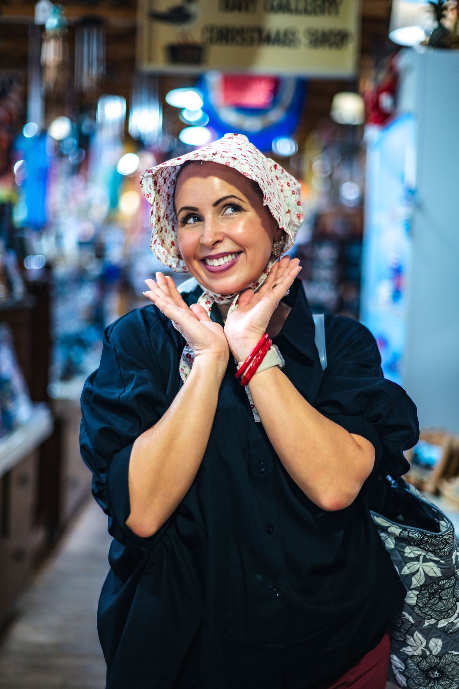 A figure in traditional period attire with a white lace bonnet smiles joyfully while posing in what appears to be a shop.
