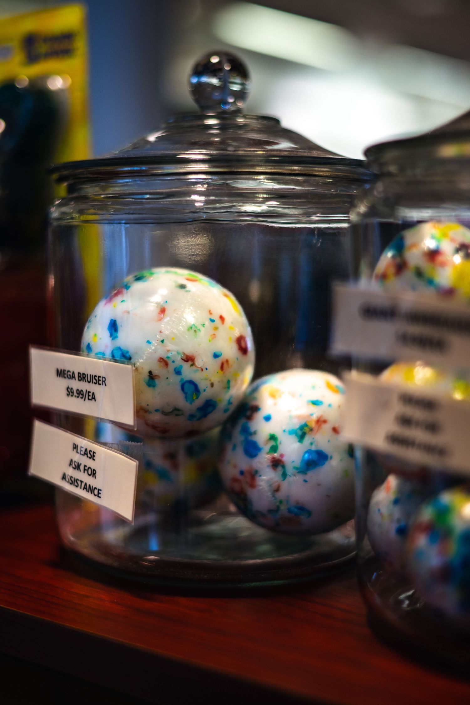 Colorful marbled bath bombs displayed in a clear glass apothecary jar with a lid on a wooden shelf.