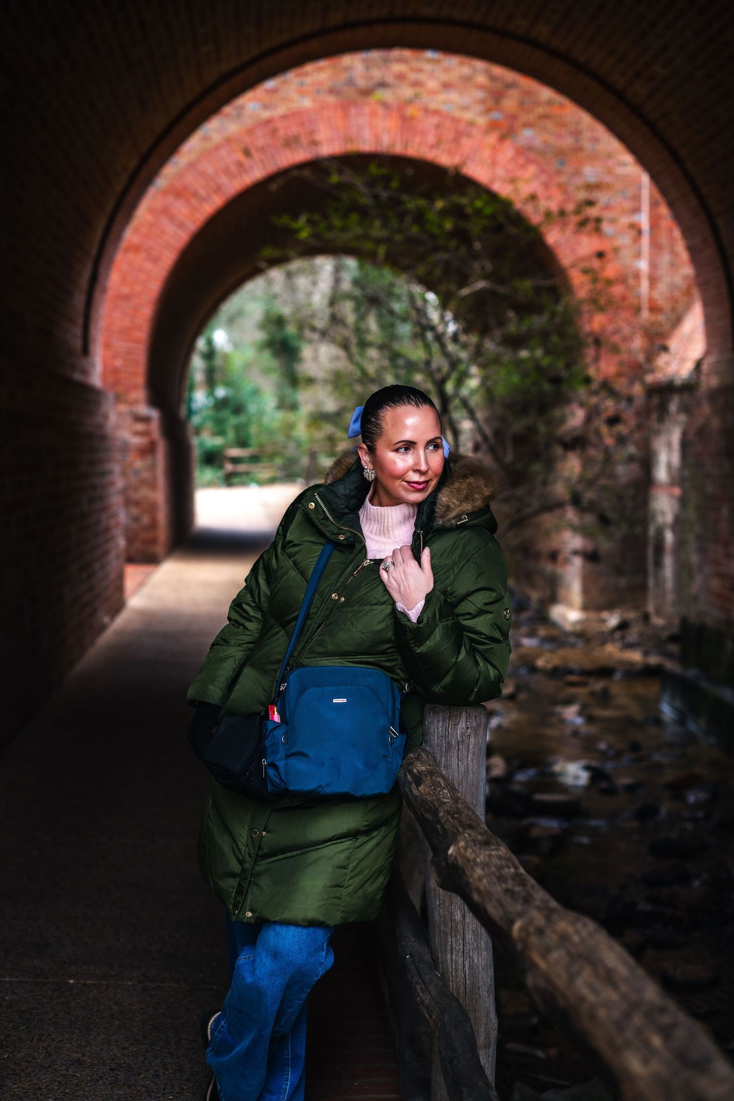 A person in a green puffer coat and blue jeans poses under a brick archway tunnel with a blue backpack.