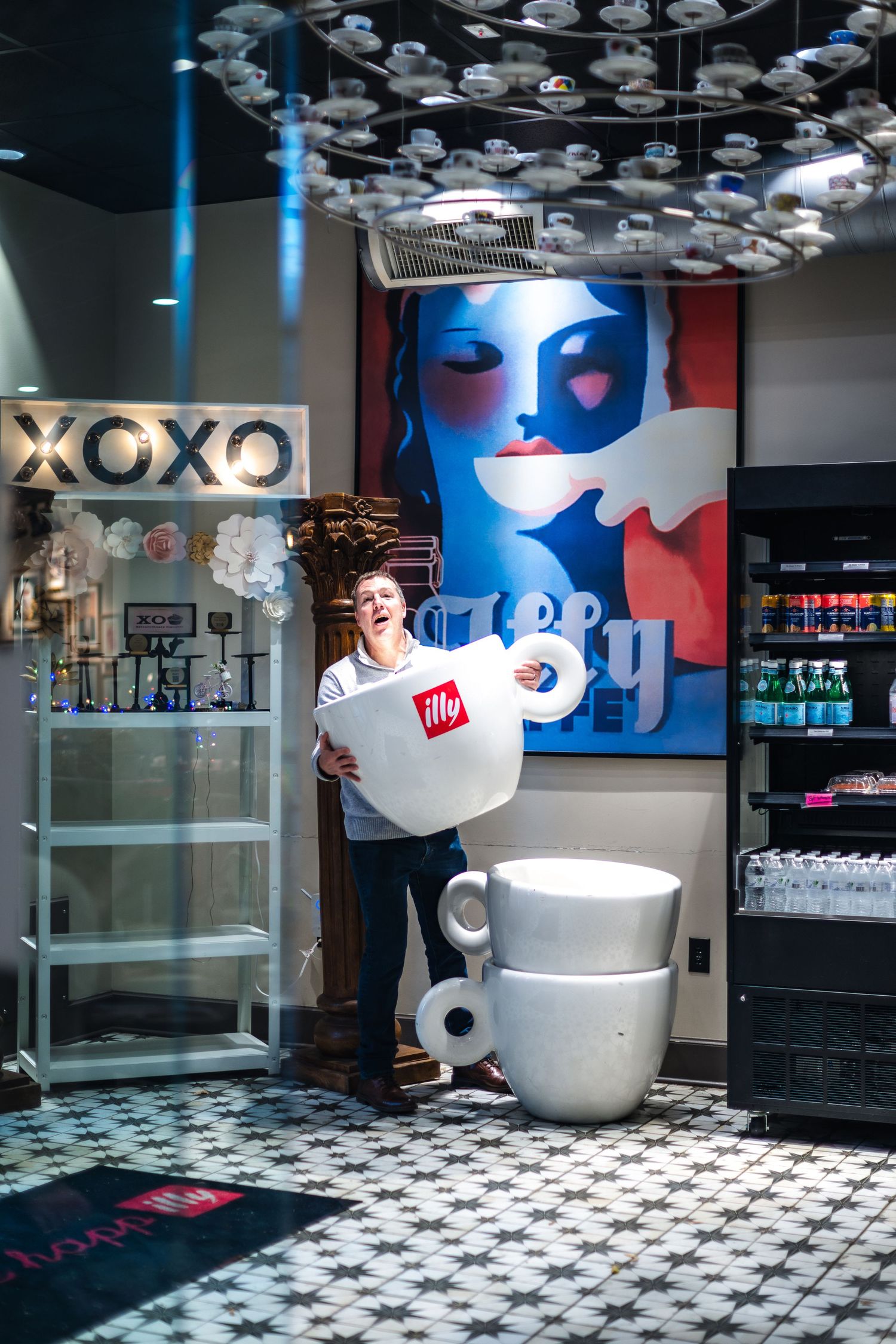Modern coffee shop interior with checkerboard floor, hanging cloud lights, and large coffee cup decor at XOXO cafe.