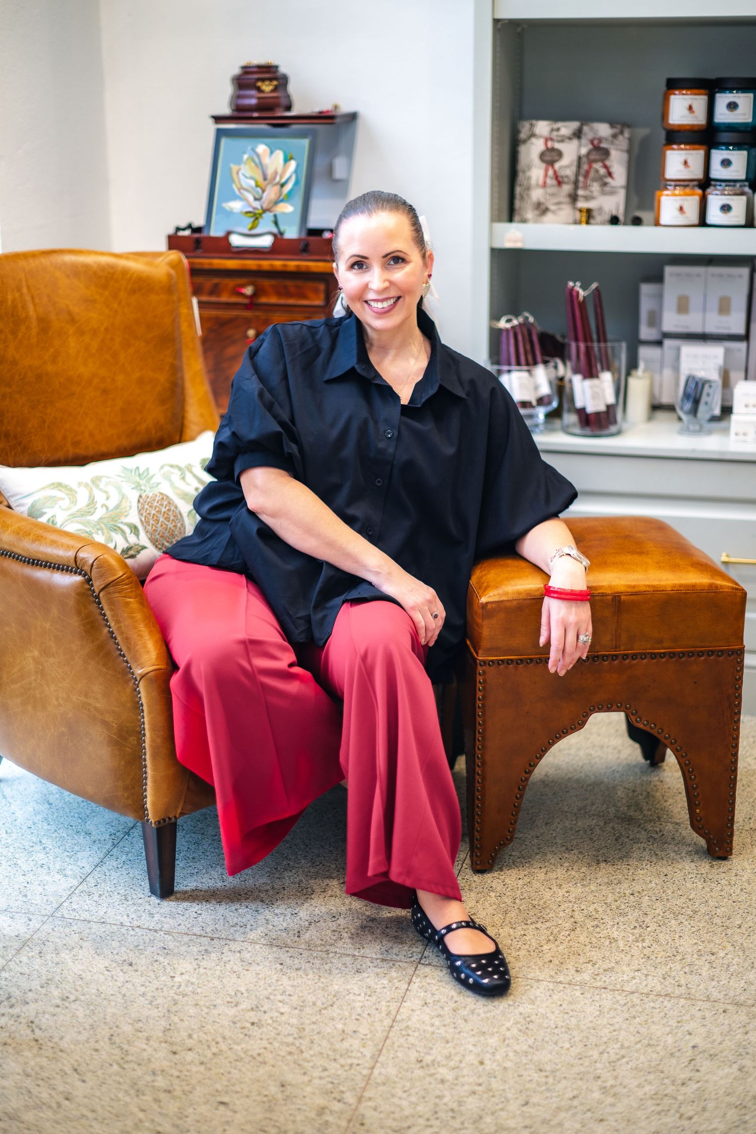 Professional wearing black button-down shirt with red pants sits casually on brown leather armchair in cozy office space.
