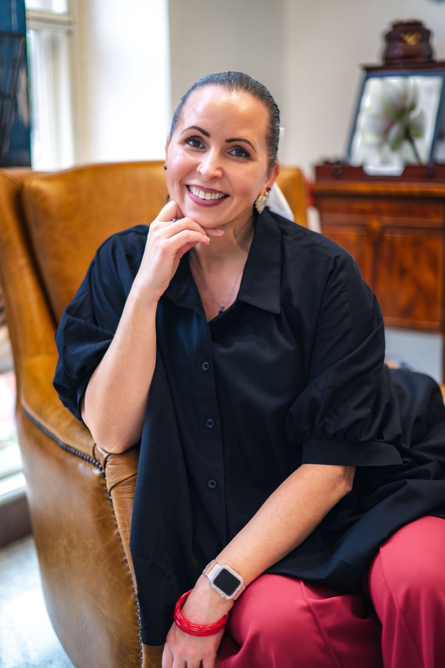 Professional in a black button-down shirt relaxes in a camel leather chair while wearing red pants and a smartwatch.